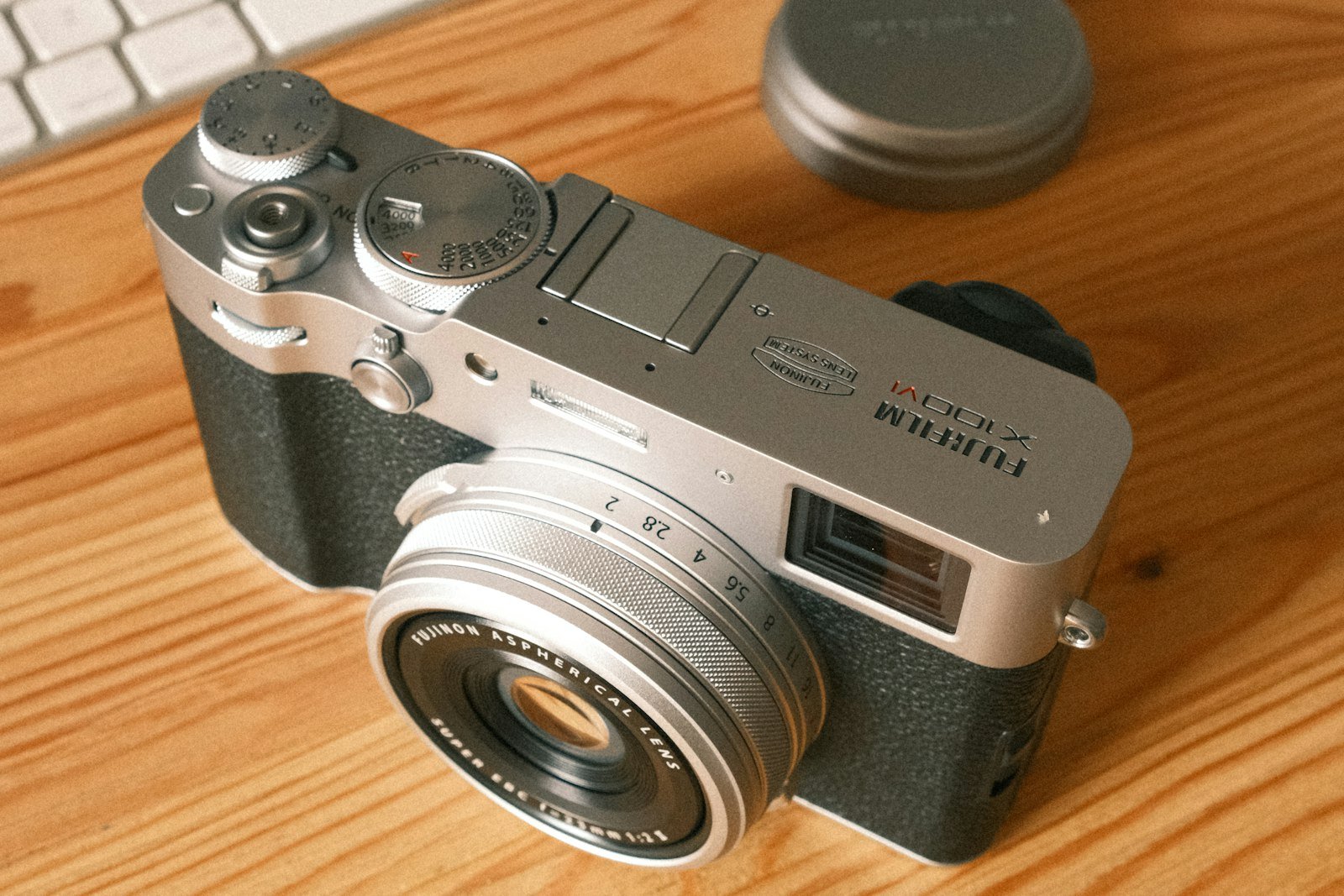 Cámara Fujifilm X100VI plateada sobre una mesa de madera, junto a un teclado y la tapa del objetivo