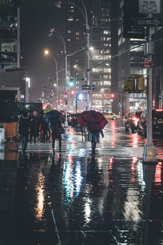 Peatones con paraguas cruzando una calle mojada de Nueva York de noche, con reflejos de luces